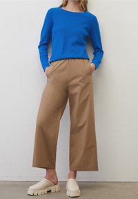 Donna che indossa un top blu a maniche lunghe, pantaloni larghi beige chiaro e scarpe slip-on chunky color avorio contro un muro neutro.
