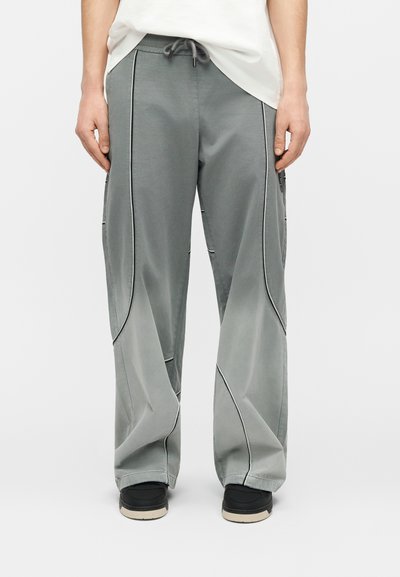 Άτομο που φοράει άνετο γκρι παντελόνι με λεπτομέρειες μαύρης τρέσας, συνδυασμένο με μαύρα παπούτσια και λευκό τοπ, στέκεται μπροστά σε λευκό φόντο.