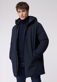 Marineblaue isolierte Parka mit Kapuze, ausgestattet mit einem Frontreißverschluss, seitlichen Taschen und glatter Stofftextur. Darunter ein marineblauer Pullover mit Zopfmuster.