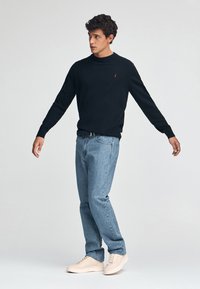 Pull noir à col rond avec logo rouge, jean en denim bleu clair et baskets beiges. Le modèle se tient avec les bras légèrement écartés.