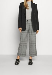 Kobieta w czarnym żakiecie, jasnoszarym golfie, szerokich spodniach w szarym kratę oraz czarnych loafersach z srebrną klamrą.