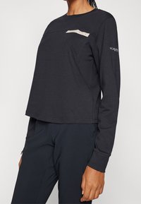 Black long-sleeve shirt made of cotton-blend material. Features a front pocket with a light tan accent and a round neckline.