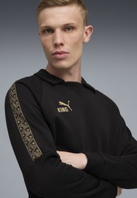 Un jeune homme aux cheveux blonds courts portant un hoodie noir avec le texte doré "KING" et des détails de manches motifs, croise les bras.