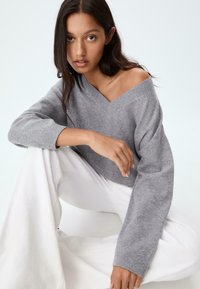 Kvinde med langt mørkt hår sidder med korslagte ben iført en grå off-shoulder sweater og hvide vidde bukser foran en lys baggrund.