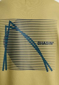 Tissu jaune avec un motif géométrique composé de lignes horizontales noires et de formes angulaires bleues, à côté du mot « CHASIN' » en texte noir.