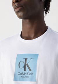 Artimo plano nuotrauka, kurioje matyti žmogus vilkintis baltą marškinėlį su mėlyno kvadrato logotipu, kuriame parašyta "cK Calvin Klein NEW YORK" ant krūtinės.