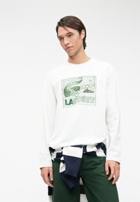 Maglietta bianca a maniche lunghe con grafica verde raffigurante un coccodrillo e la scritta 'LA COTE'; abbinata a un maglione a righe navy e bianche legato in vita.