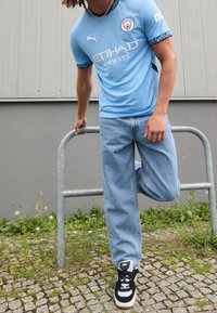 Taivaansininen Manchester City -pelipaita mustilla yksityiskohdilla, valkoisella logolla ja tekstillä "ETIHAD AIRWAYS". Yhdistetty vaaleansinisiin farkkuihin ja mustiin lenkkareihin.