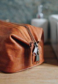 Brown leather toiletry bag with a zipper closure, featuring a smooth texture and visible stitching details. Shaped rectangular with rounded edges.