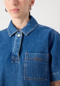 Blå denimskjorte med krage, to-knapps placket, brystlomme og kontrasterende oransje sømdetaljer. Glatt tekstur.