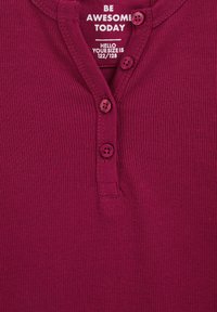 Maglietta henley a coste color bordeaux con placca a bottoni e tre bottoni viola. Include testo bianco stampato sul colletto e sull'etichetta della taglia.