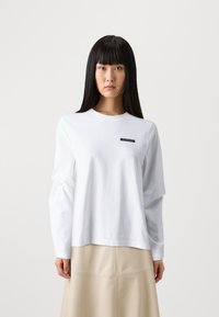 White long-sleeve t-shirt in cotton with a round neckline, featuring a black logo patch on the left chest area. Smooth texture and relaxed fit.