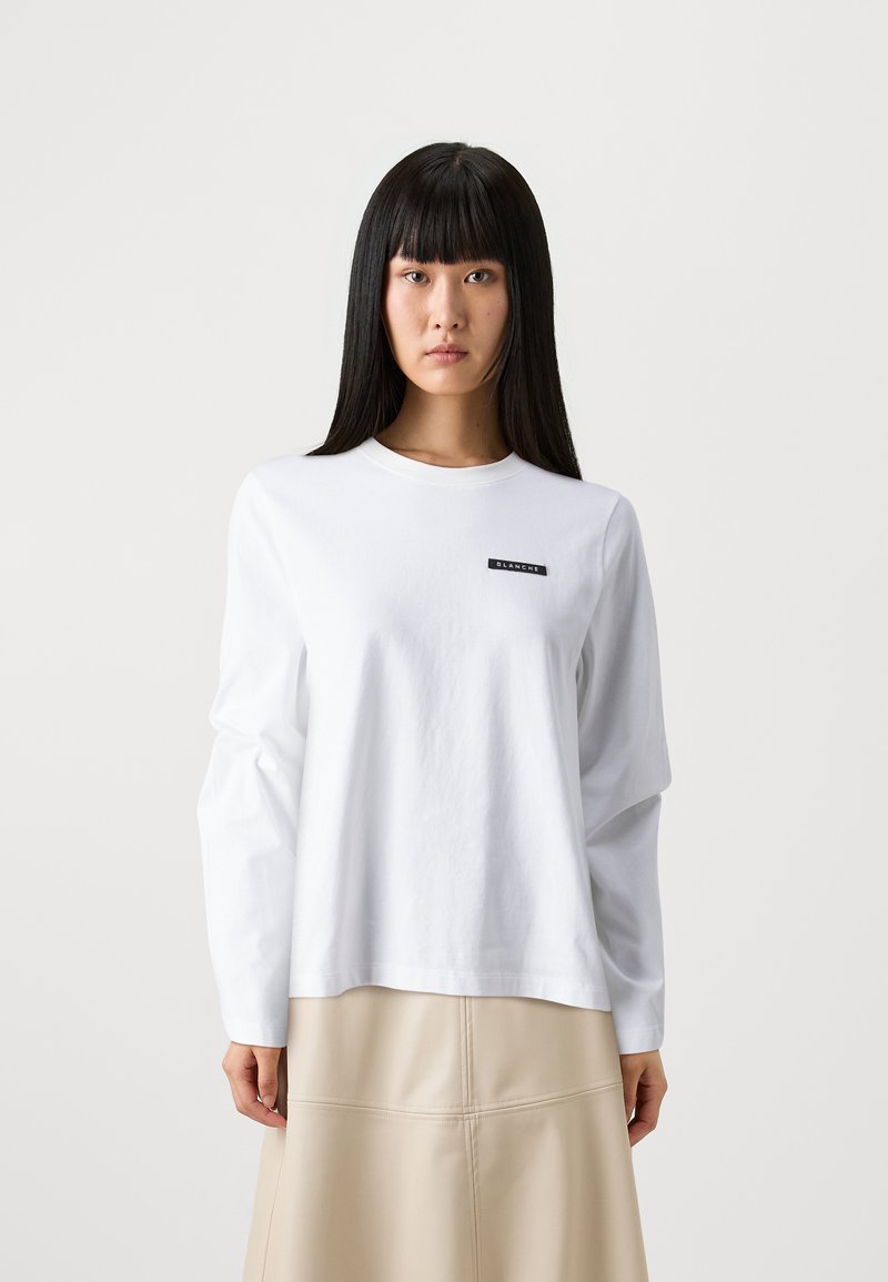 White long-sleeve t-shirt in cotton with a round neckline, featuring a black logo patch on the left chest area. Smooth texture and relaxed fit.