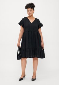 Black dress with short ruffled sleeves, V-neck, dotted texture, tiered skirt, paired with a black bag and black pointy-toed heels.