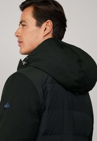 Muškarac u crnoj jakni s punjenjem i kapuljačom, okrenut lijevo, prikazuje bočni profil i gornji dio leđa s vidljivim logotipom marke na rukavu.