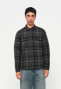 Young man wearing a black and gray checkered button-up jacket with chest pockets and faded blue jeans against a plain white background.