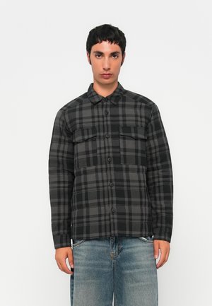 Young man wearing a black and gray checkered button-up jacket with chest pockets and faded blue jeans against a plain white background.