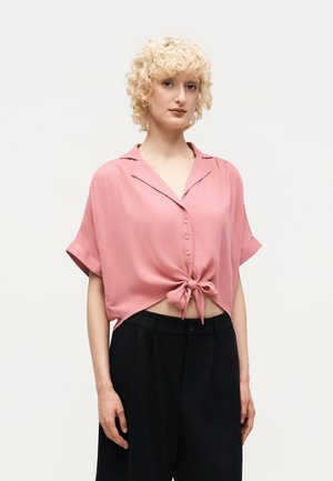 Άτομο με κοντά, σγουρά ξανθά μαλλιά, φορούν ροζ κοντομάνικο πουκάμισο με κουμπιά δεμένο στη μέση και μαύρο παντελόνι με ψηλή μέση.