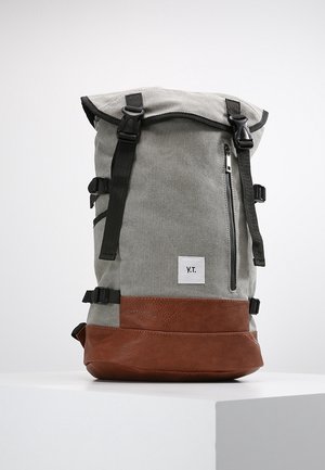 Sac à dos en toile grise avec fond en cuir marron, bretelles réglables, poche avant zippée et une étiquette blanche avec "Y.T." sur le côté.