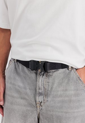 Cintura in nylon nero con fibbia di plastica, caratterizzata da una superficie strutturata, indossata con jeans di denim grigio chiaro e una t-shirt bianca.