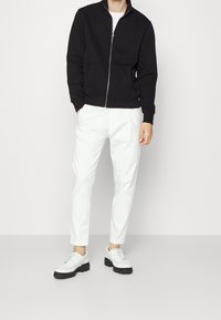 Svart zip-up sweatshirt med två framfickor, kombinerad med vita avsmalnande byxor och vita sneakers med svarta sulor.