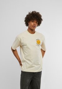 T-shirt en coton crème avec un col rond et des manches courtes. Présente un visage souriant jaune imprimé et du texte sur la zone du côté gauche de la poitrine.