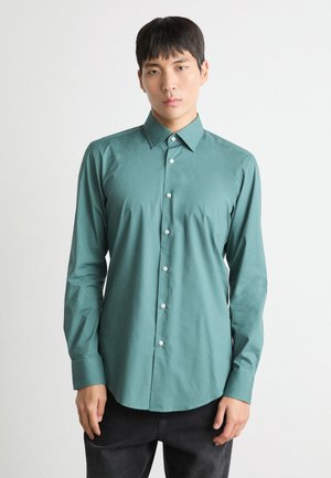 Camicia a maniche lunghe in teal realizzata in cotone, con colletto button-down, bottoni frontali e una consistenza liscia. Abbinata a pantaloni neri.