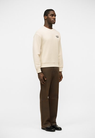 Άνδρας που στέκεται φορώντας κρεμ φούτερ με μικρό κείμενο στο στήθος, καφέ ίσια παντελόνια και μαύρα δερμάτινα παπούτσια, σε ένα απλό λευκό φόντο.