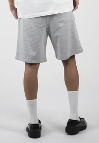 Personne portant un short gris clair jusqu'aux genoux, des chaussettes blanches côtelées, des mocassins noirs brillants et une chemise blanche, debout devant un fond uni.