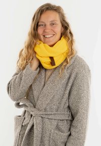 Geel gebreide infinity sjaal met een textuurpatroon, voorzien van een bruine leren label, gedragen over een grijze, pluizige badjas.