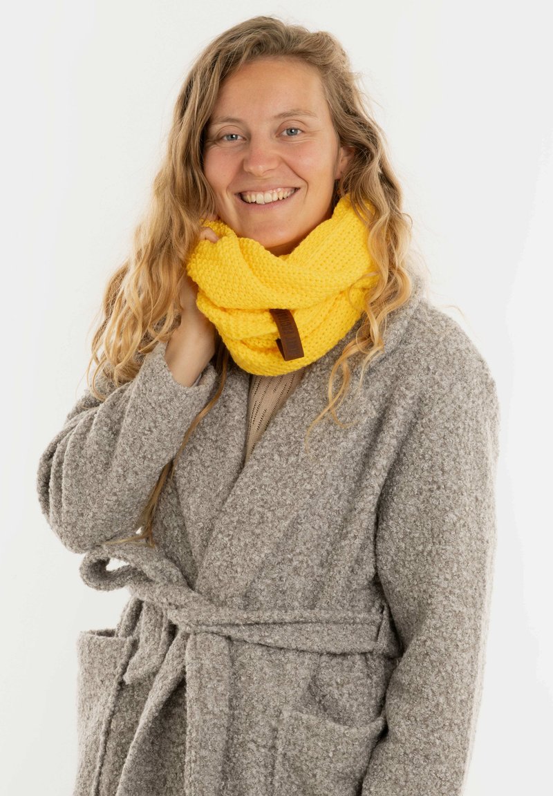 Geel gebreide infinity sjaal met een textuurpatroon, voorzien van een bruine leren label, gedragen over een grijze, pluizige badjas.