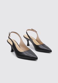 Tacones slingback de cuero negro con punta afilada, un tacón moderado y un diseño limpio. Presenta una delgada correa en el tobillo con una hebilla en tono dorado.