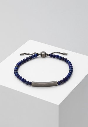Bracelet ajustable bleu à perles avec fermoir en métal foncé et plaque métallique rectangulaire gravée sur une surface blanche.