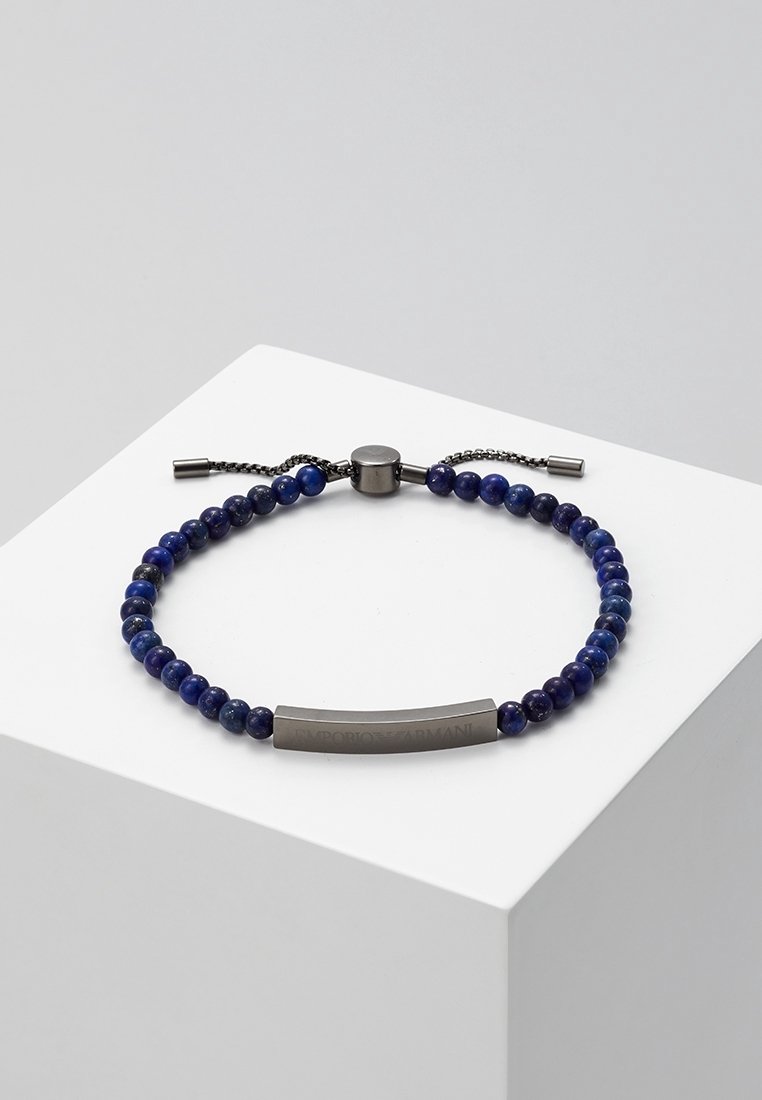 Bracelet ajustable bleu à perles avec fermoir en métal foncé et plaque métallique rectangulaire gravée sur une surface blanche.