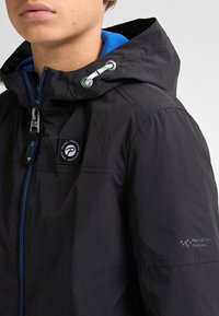 Jachetă impermeabilă neagră cu interior albastru, închidere cu fermoar, glugă ajustabilă și patch cu logo pe piept. Țesătură netedă, cu textură minimă.