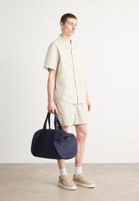 Navy duffel bag with dual handles, zipper closure, and a round shape. Model wears light striped short-sleeve shirt and shorts. Sneakers and white socks.