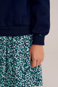 Temno modra majica s kapuco z rebrastimi manšetami, v paru z nabrano krilom z zeleno-belimi cvetličnimi vzorci na temno modri podlagi.