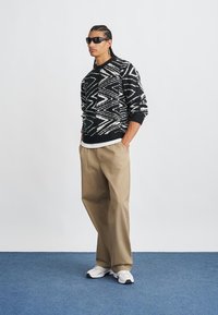 Schwarz-weißer gemusterter Pullover mit Rundhalsausschnitt, kombiniert mit beigen weiten Hosen und weißen Turnschuhen. Model trägt eine Sonnenbrille.