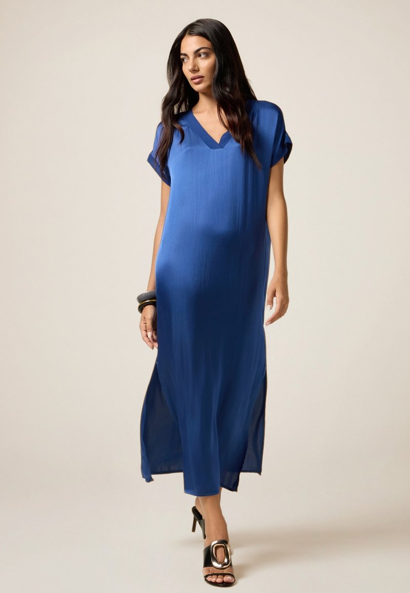 Donna che cammina in un vestito blu lungo fino alla caviglia, morbido, con scollatura a V e spacchi laterali, indossando sandali con tacco nero e braccialetti a un polso.