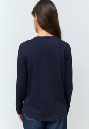 Kobieta z długimi brązowymi włosami, nosząca małe kolczyki-koła, granatową bluzkę z długim rękawem i niebieskie dżinsy, zwrócona tyłem na jednolitym tle.