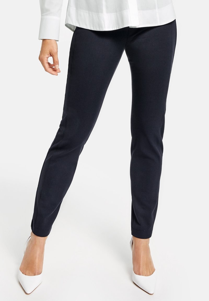 Femme portant un pantalon noir ajusté, une chemise blanche boutonnée et des escarpins blancs à bout pointu, debout devant un fond uni.