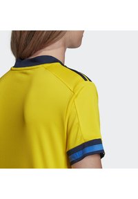 Gul sportstrøje med tekstureret stof, der har en marineblå krave og ærmekanter med blå striber. Nærbillede set fra bagsiden.