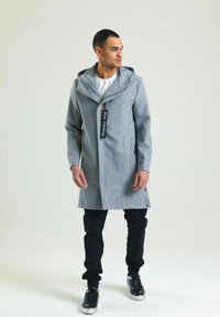 Grauer langer Mantel mit hohem Kragen, Reißverschluss und zwei Seitentaschen, kombiniert mit schwarzen Hosen und glänzenden schwarzen Sneakers.