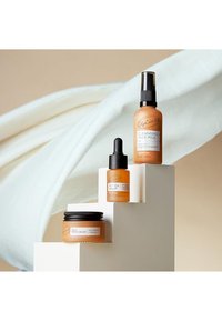 Drei Hautpflegeprodukte auf weißen Podesten: ein braunes Glas, eine Tropfflasche und eine Sprühflasche, alle mit oranger Verpackung.