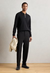 Polo negro de manga larga con cremallera plateada, pantalones negros de corte ajustado, zapatos negros y una chaqueta beige clara colgada del brazo.