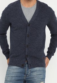 Navy knitted cardigan with a v-neckline, textured pattern, five front buttons, and ribbed cuffs, paired with a grey shirt and denim jeans.