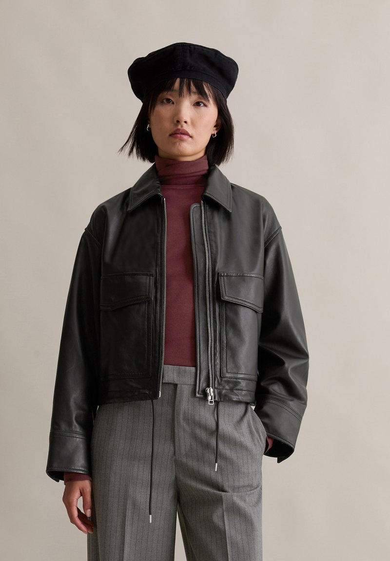 Schwarze Lederjacke mit Reißverschluss vorne, zwei vorderen Taschen und Kragen, kombiniert mit einem kastanienbraunen Rollkragenpullover und grauen Nadelstreifenhosen.