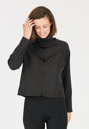 Smilende kvinde med kort brunt hår, iført en sort lynlåsjakke og sorte leggings, rører ved sit hår med den ene hånd.