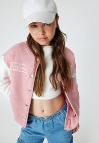 Roze en witte bomberjack met drukknoopsluitingen, voorzien van borduurwerk "Los Angeles". Gemaakt over een witte ribbelige crop top en blauwe sweatpants met trekkoord.