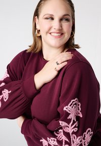 Bordeauxrode trui met ballonmouwen, voorzien van roze bloemenborduursel met pailletten op de mouwen. Model draagt creolen en ringen.